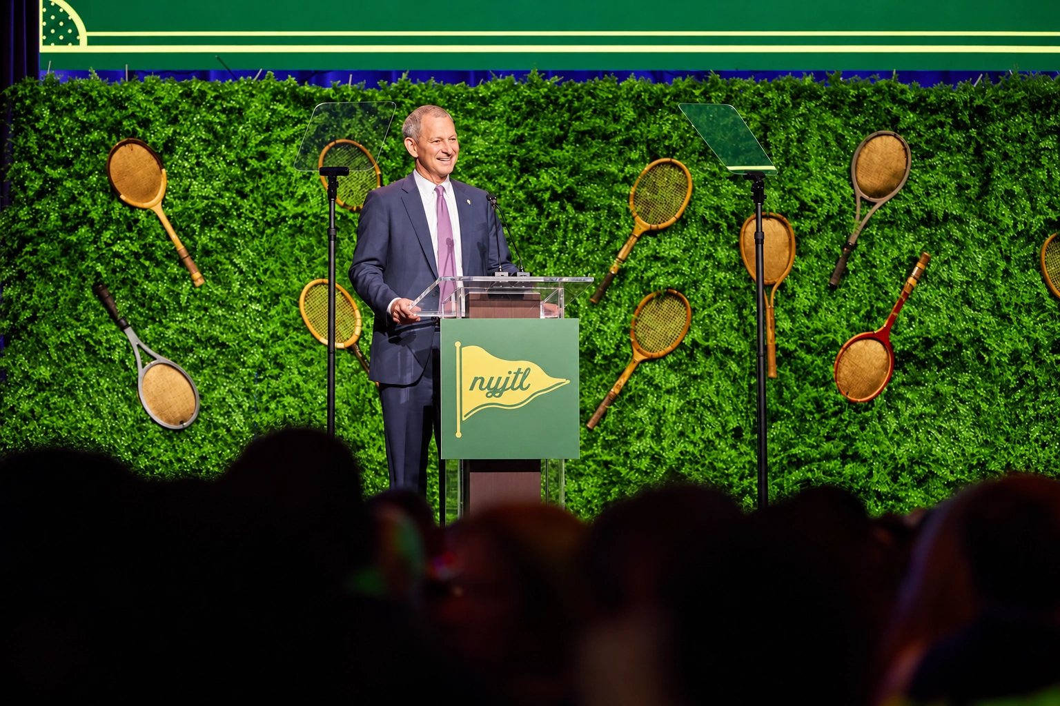 Wide photograph of a speaker at the NYJTL podium, with tennis racquets behind him and audience silhouettes in the foreground